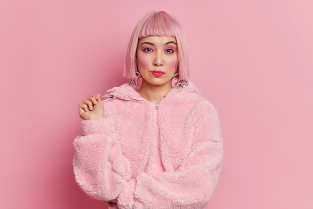 serious pretty asian woman with trendy pink hair dressed in winter coat has bright vivid makeup poses 