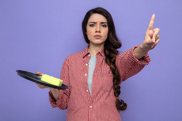 Serious pretty caucasian cleaner woman holding sponge on plate and pointing up