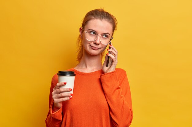 Serious pretty European girl has boring telephone conversation, hangs out sometime, talks to person, holds disposable coffee, uses modern technologies, dressed in sweater, poses over yellow wall