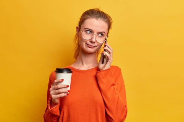 Серьезная симпатичная европейская девушка ведет скучный телефонный разговор, иногда тусуется, разговаривает с человеком, держит одноразовый кофе, использует современные технологии, одета в свитер, позирует у желтой стены