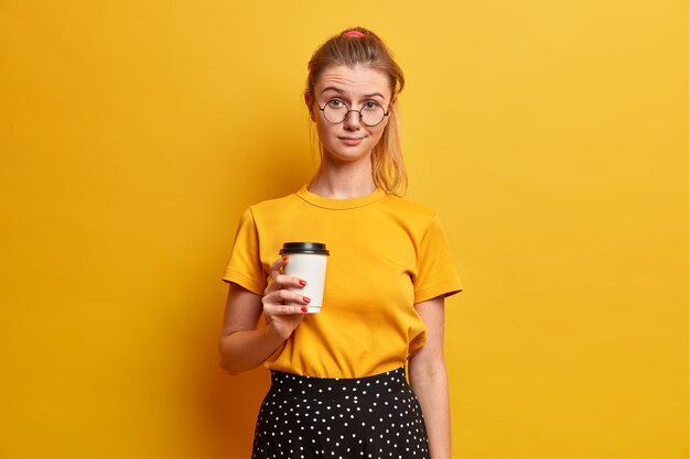 진지한 예쁜 여자가 새로운 친구와 자유 시간을 보내고 테이크 아웃 커피 음료 음료를 들고 불행하게 보이는 노란색 티셔츠 광학 안경 스탠드 실내