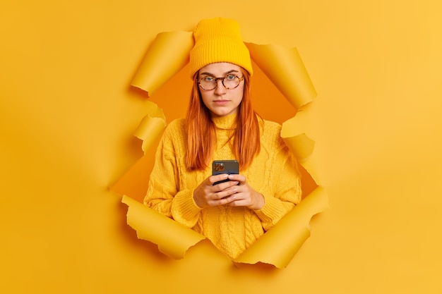 Serious pretty redhead woman using smartphone, wears yellow hat and sweater, breaks through paper wall