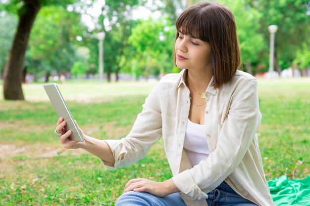 Serious pretty woman reading news on tablet and sitting on lawn