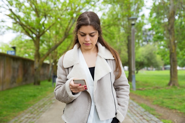 Serious pretty young woman using smartphone in park