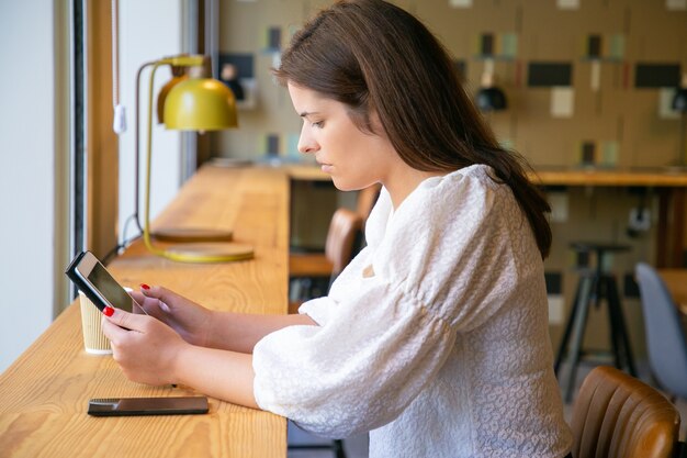 Serious pretty young woman in white shirt using tablet while sitting at desk in co-working space or coffee shop