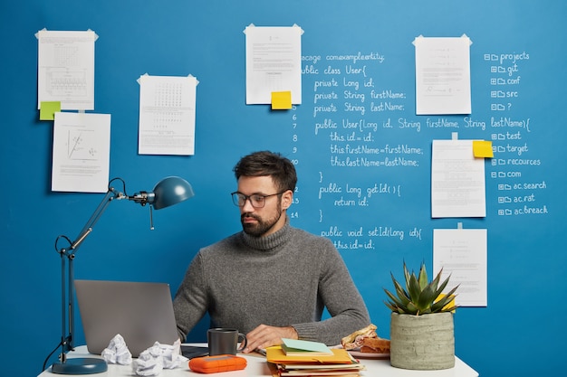 Serious professional male geek concentrated at monitor of modern laptop, wears optical glasses, poses in coworking space against blue background