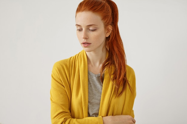 serious red-haired female with freckled skin with no make-up, wearing yellow cape, standing crossed hands. Beautiful woman with reddish pony tail, looking down