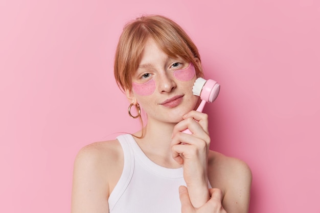 Serious redhaired millennial girl uses face massager applies hydrogel patches under eyes wears casual white t shirt isolated over pink studio background Skin care and facial treatments concept