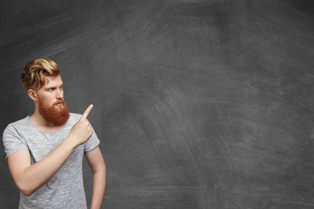 Serious redhead bearded Caucasian hipster student dressed in gray t-shirt standing in classroom pointing at blank copy space wall with his finger, showing something on it.