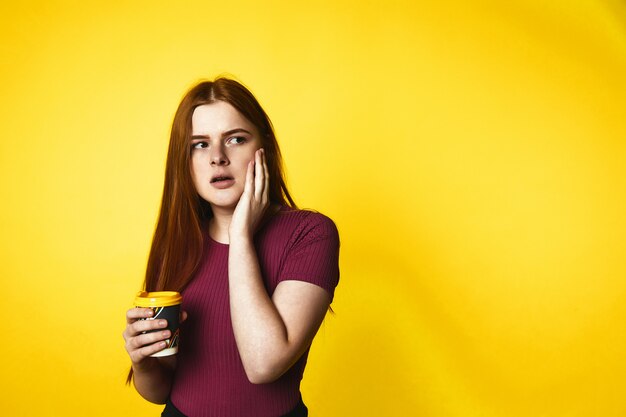 Serious redhead caucasian girl is holding drink in one hand and holding cheek with another