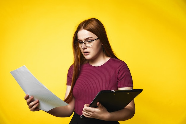 Serious redhead caucasian girl is holding folder with documents in hands with surprised face