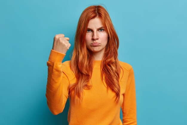 Серьезная рыжая европейка гневно смотрит, показывает кулак, просит не трогать кошельки губы и с раздраженным выражением лица одетая в повседневный оранжевый джемпер.