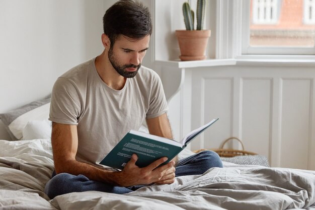Serious relaxed unshaven man holds book in front of face, dressed in casual t shirt, sits in lotus pose on bed
