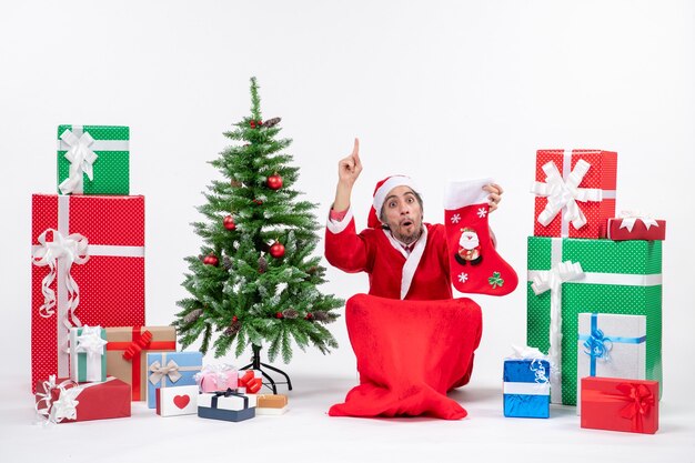 Serious santa claus pointing above sitting on the ground and holding christmas sock near gifts and decorated new year tree on white background