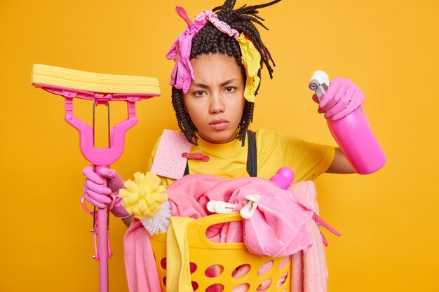 Serious scrupulous Afro American housewife has dreadlocks holds with cleaning accessories