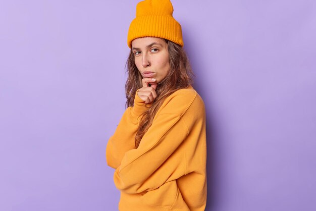 Serious self confident beautiful teenage girl holds chin looks directly at camera considers something stands sideways against purple wears stylish hat and jumper. Human face expressions