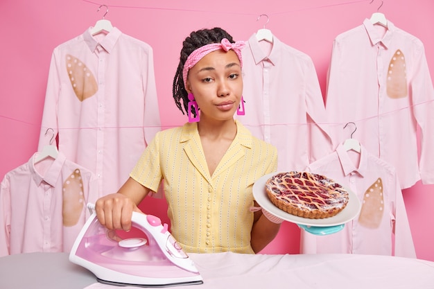 Serious self confident housewife wears headband domestic clothes irons laundry bakes delicious pie poses in dressing gown. Housekeeping process