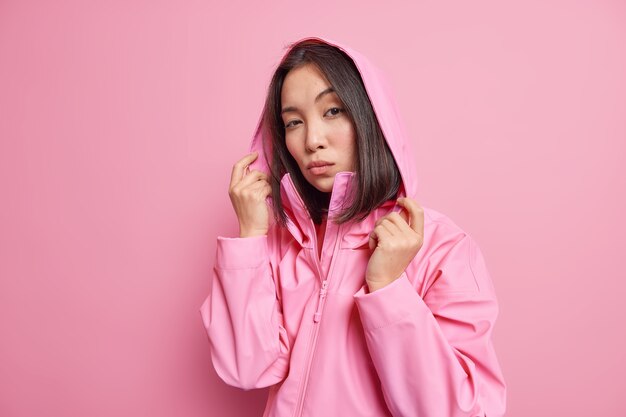 Serious self confident millennial girl with dark hair wears anorak hood on head looks directly  models against pink wall going to have walk during cold windy day. People and style
