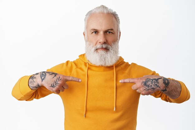 Serious senior guy with tattoos and hipster beard, pointing fingers at himself, point at his hoodie, looking at front, standing against white wall.