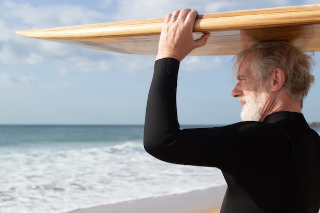 Free photo serious senior man holding surfboard on head. gray-haired bearded man in wetsuit with wooden board on head looking and enjoying sea and waves views. active and healthy lifestyle of aged people concept