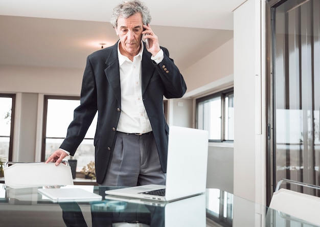 Serious senior man talking on mobile phone with laptop and digital tablet on reflective desk