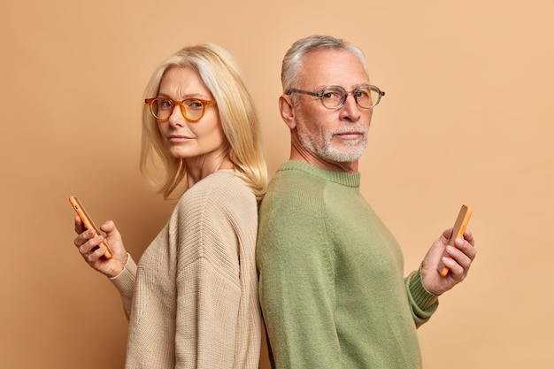 Free photo serious senior woman and her husband hold modern gadgets read media spend free time in internet ignore each other stand backs wear spectacles sweater isolated over beige wall