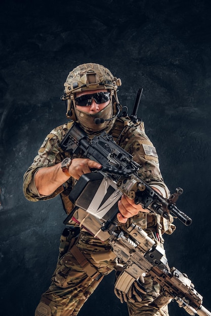 Free photo serious soldier in full gear and military uniform is standing with machine gun over dark background.