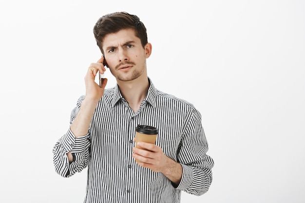 Serious strict caucasian bearded brother in striped shirt, holding cup of coffee and talking on phone with focused expression, feeling intense discussing important business meeting over gray wall
