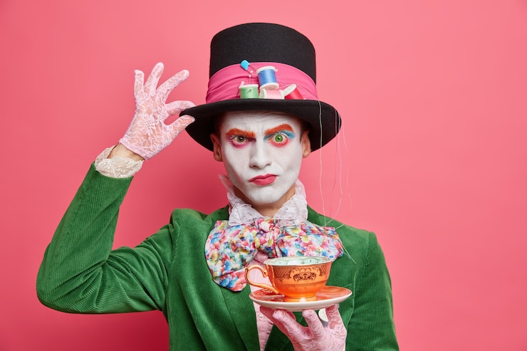 Free Photo serious strict male hatter keeps hand on tall hat holds cup of tea poses on halloween carnival has bright professional makeup poses over rosy wall