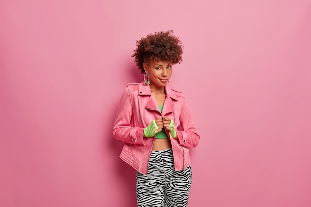 Una seria ed elegante donna afroamericana indossa una giacca rosa alla moda, guanti sportivi e leggings, sembra sicura di sé, ha un'acconciatura riccia