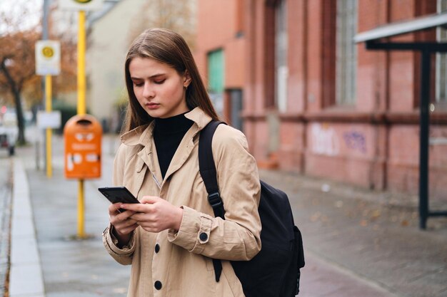 Серьезная стильная девушка с рюкзаком с помощью мобильного телефона ждет общественный транспорт на автобусной остановке под открытым небом