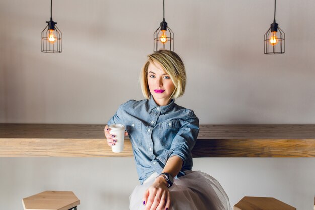 금발 머리와 분홍색 입술이 나무 의자와 테이블 커피 숍에 앉아 심각한 세련된 소녀. 그녀는 cofee 한 잔을 들고