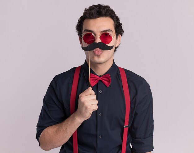 Serious stylish man with bow tie wearing glasses and suspenders looking at front holding mustache on stick standing over white wall