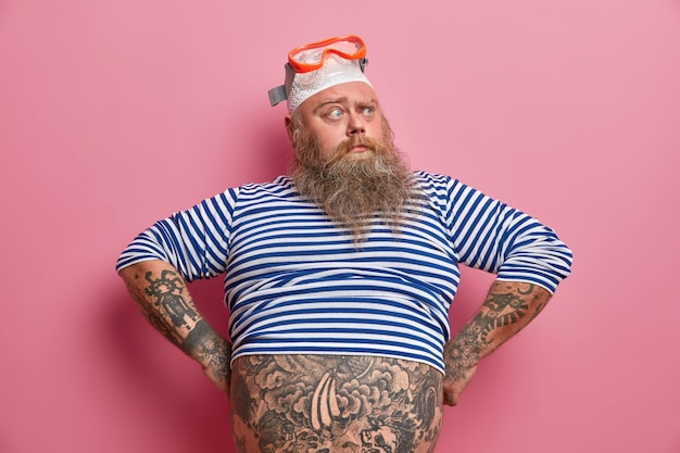 Free photo serious surprised man diver wears swimming goggles on head and striped sailor jumper, keeps hands on hips, confused to hear something strange, has tattooed belly, isolated on pink wall.