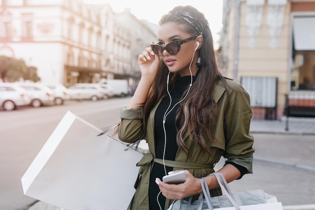Serious tanned woman in dark glasses listening music in white earphones. Charming hispanic lady in trendy jacket carrying purchases, walking on the street.