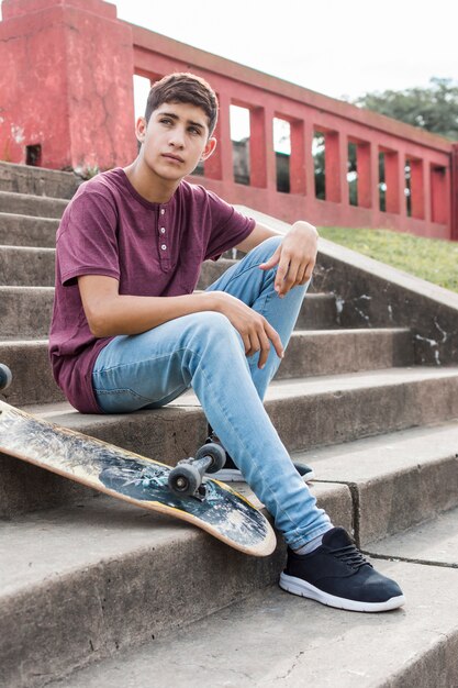 Serious teenage boy sitting on staircases with skateboard looking away