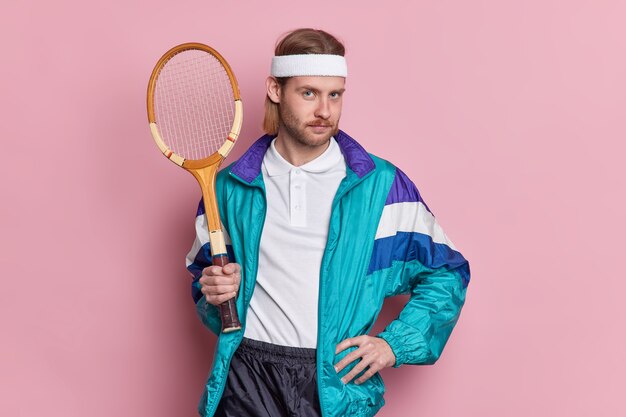 Серьезный теннисист со спортивным снаряжением держит руку на талии, выглядит уверенно готовым к игре, ведет активный образ жизни.