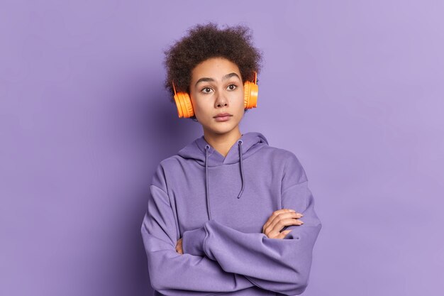 Serious thoughtful African American girl keeps hands crossed listens music thinks about something wears casual sweatshirt.