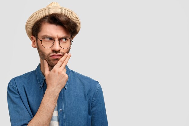 Serious thoughtful man farmer holds chin, thinks how to succeed in agricultural sphere, dressed in stylish straw hat and casual shirt