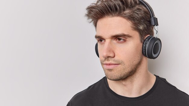 Serious thoughtful man with dark hair concentrated away listens educational content via headphones poses against white background blank copy space for your advertising content Deep thoughts