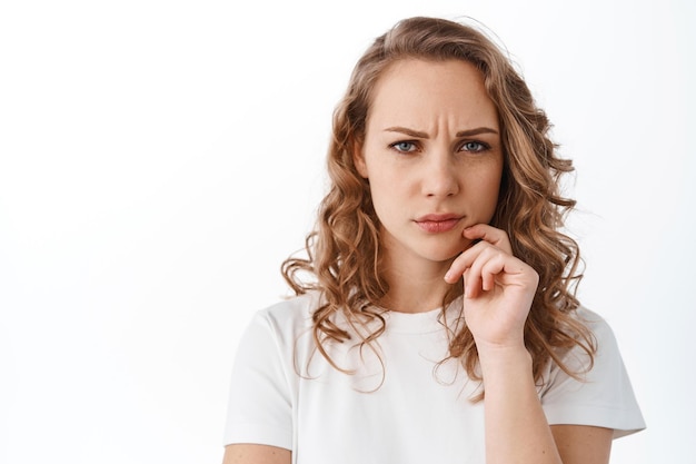 Serious and thoughtful woman frowns and looks at camera pensive, thinking, standing over white background