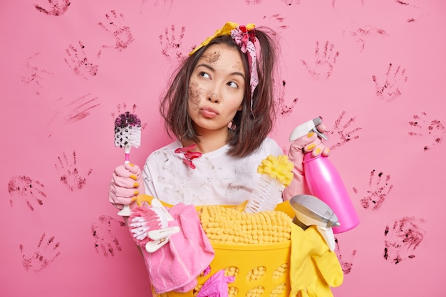 Free photo serious thoughtful young asian housewife holds detergent and brush looks pensively has dirty face poses near basket of laundry isolated over pink wall