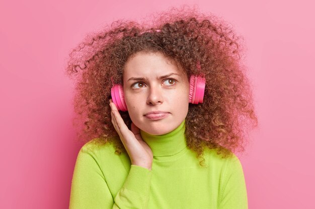 Serious thoughtful young European woman with curly bushy hair wears wireless stereo headphones on ears smirks face has dissatisfied expression wears green poloneck isolated over pink bacground