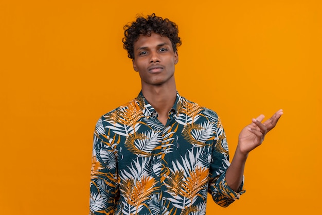 Серьезный и вдумчивый молодой красивый темнокожий мужчина с вьющимися волосами в рубашке с принтом листьев смотрит в камеру, поднимая руку на оранжевом фоне