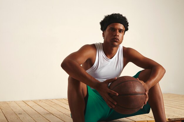 休憩を取っている彼の手でボールを持って床に座っている深刻な疲れたバスケットボール選手