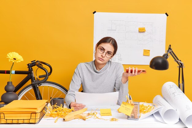 Serious tired woman holds paper sketch and smartphone poses at desktop prepares for brainstorming meeting or briefing with colleagues almost finished engineering project works on blueprints.