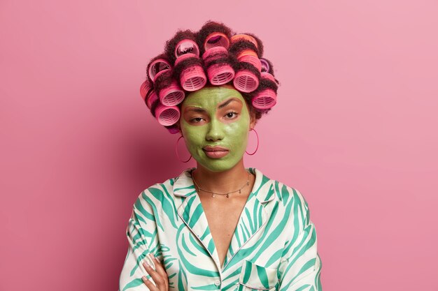 Serious unimpressed woman stands with crossed hands over body, feels bored during beauty procedures, wears hair rollers for creating perfect curls, green mask to rejuvenate and reduce wrinkles