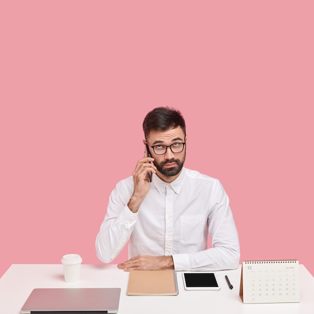 Serious unshaven intelligent man calls customer service, has telephone conversation in break of work, dressed in white shirt, focused upwards