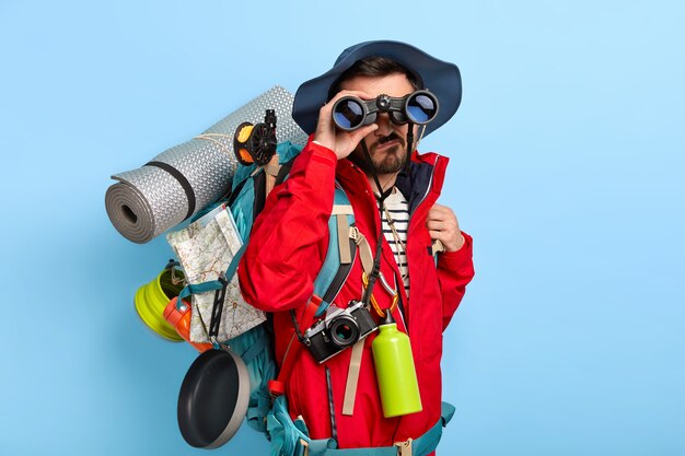 Serious unshaven male backpacker keeps binoculars near eyes, wears hat and red jacket, explores new way, carries tourist backpack