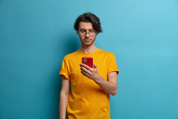 Serious unshaven man holds cell phone in hand, concentrated in screen, watches tutorial video, types answer or reads feedback about new project, wears transparent glasses, isolated on blue wall